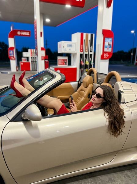 Девушка в тёмных очках полулёжа в бежевом кабриолете Ferrari, ноги в красных туфлях на стекле