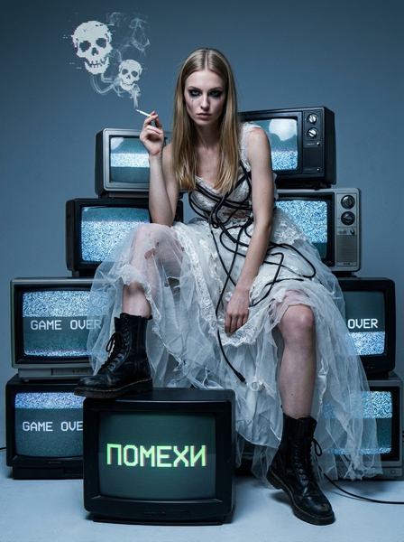 Девушка в рваном свадебном платье и ботинках сидит на пирамиде старых телевизоров с помехами